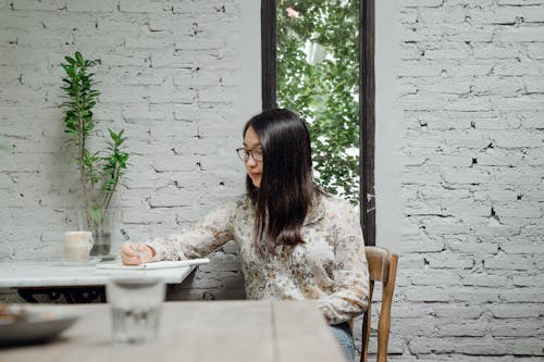 Pensive young Asian female writer in casual clothes and eyeglasses taking notes in diary while creating new article in cozy cafe