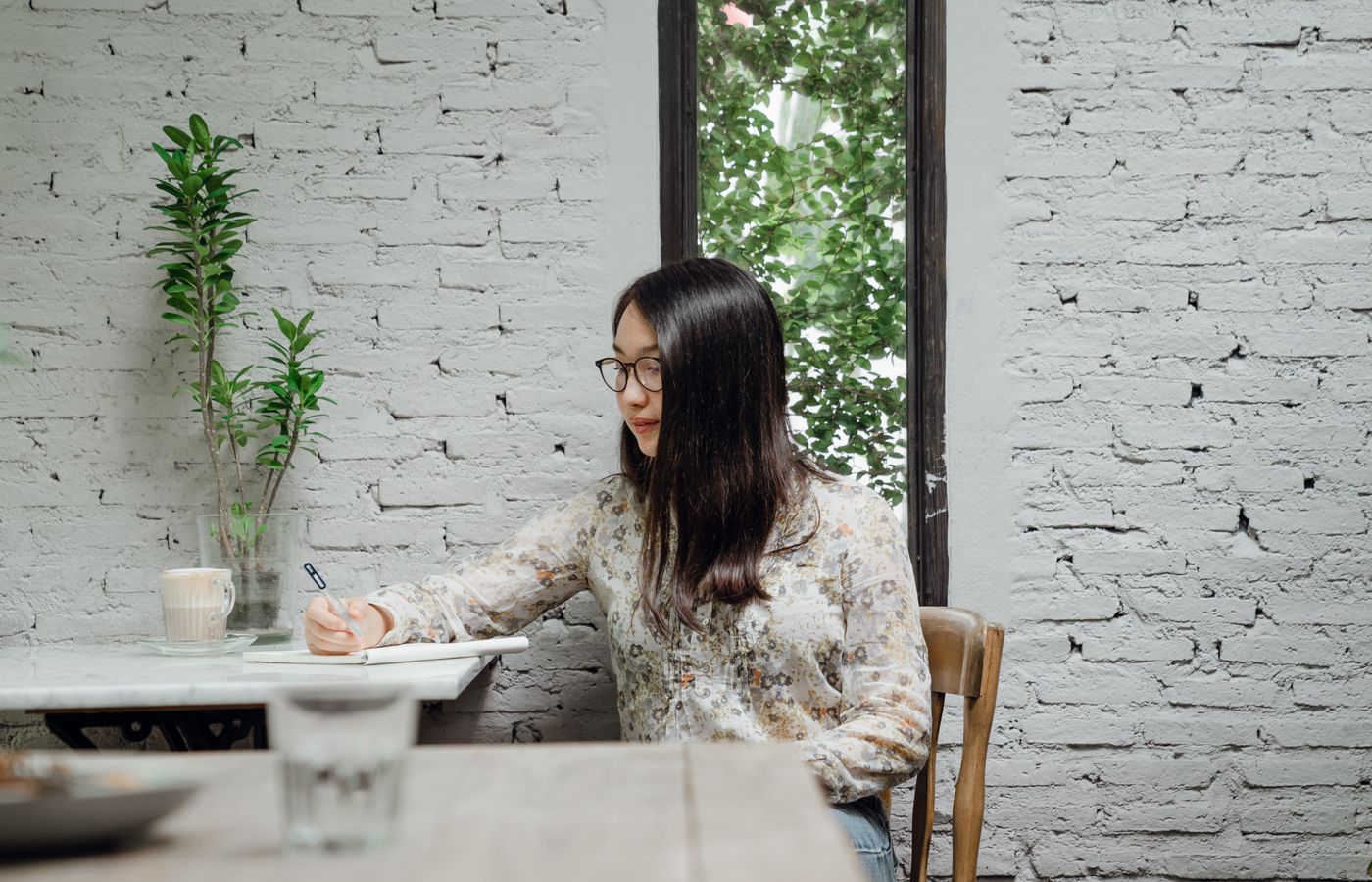 A young woman writes in a notebook, enjoying a latte in a cozy café with brick walls.