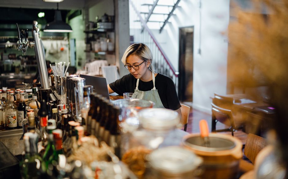 Focused young ethnic woman in apron and eyeglasses sitting at bar counter full of various drinks and using laptop