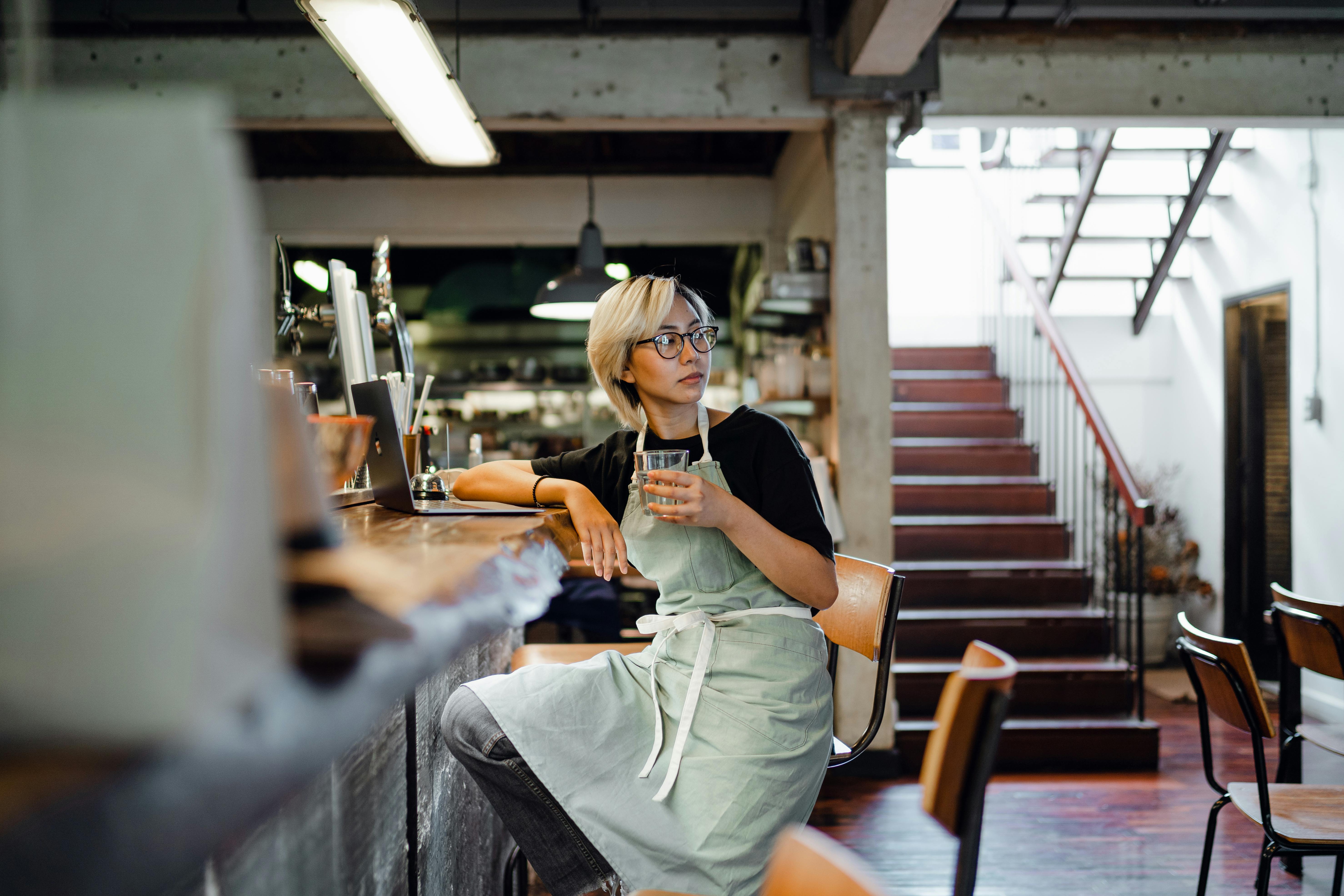 一位戴着眼镜、穿着围裙的年轻亚洲女服务员坐在柜台前，目光移开，手里拿着上网本