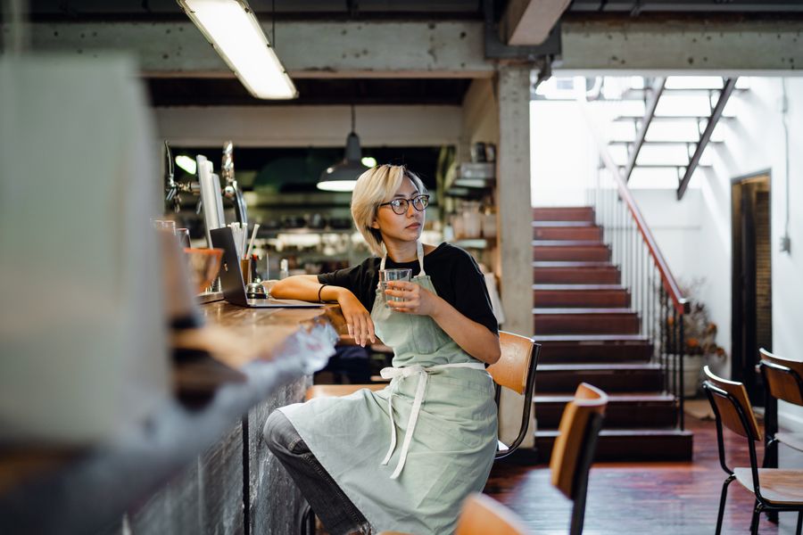 Concentrated young Asian waitress in apron and eyeglasses drinking water while sitting at counter with netbook and looking away