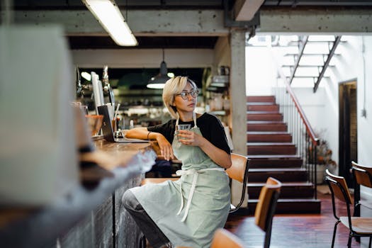 一位戴着眼镜、穿着围裙的年轻亚洲女服务员坐在柜台前，目光移开，手里拿着上网本