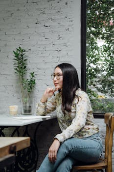 Focused young ethnic female entrepreneur in casual clothes and eyeglasses thinking about business ideas while working remotely in cozy cafe