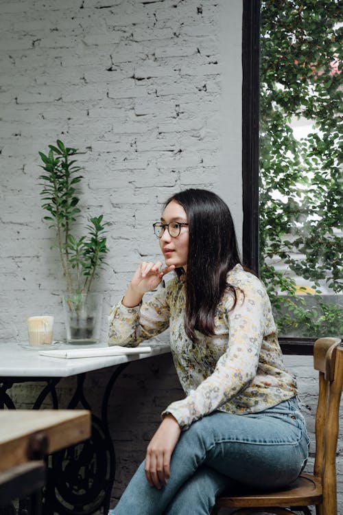Focused young ethnic female entrepreneur in casual clothes and eyeglasses thinking about business ideas while working remotely in cozy cafe