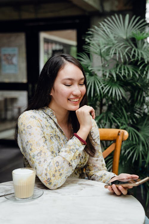 Positive young Asian female in casual clothes having video conversation while sitting in cozy cafeteria with glass of cappuccino