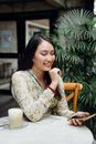 一位穿着休闲装的年轻亚洲女性正坐在舒适的自助餐厅里，一边喝着卡布奇诺一边进行视频通话