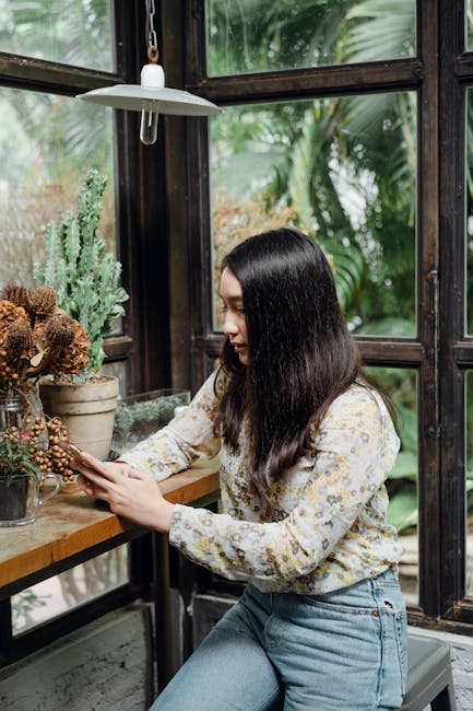 Concentrated young ethnic female in casual wear reading news on mobile phone while resting in cozy space against window with view of green plants
