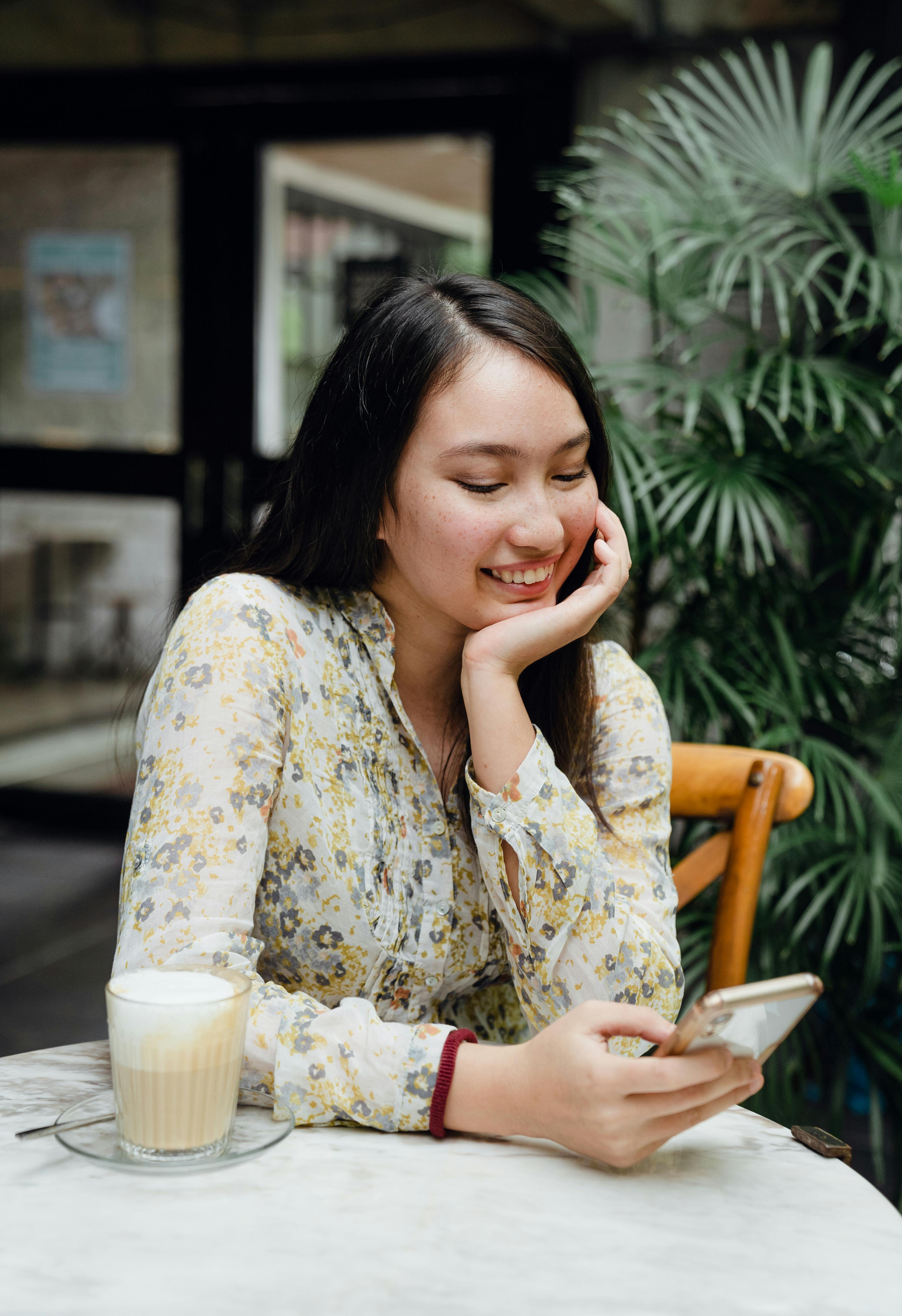 smiling young asian woman sitting in cafe with chin on hand and using smartphone