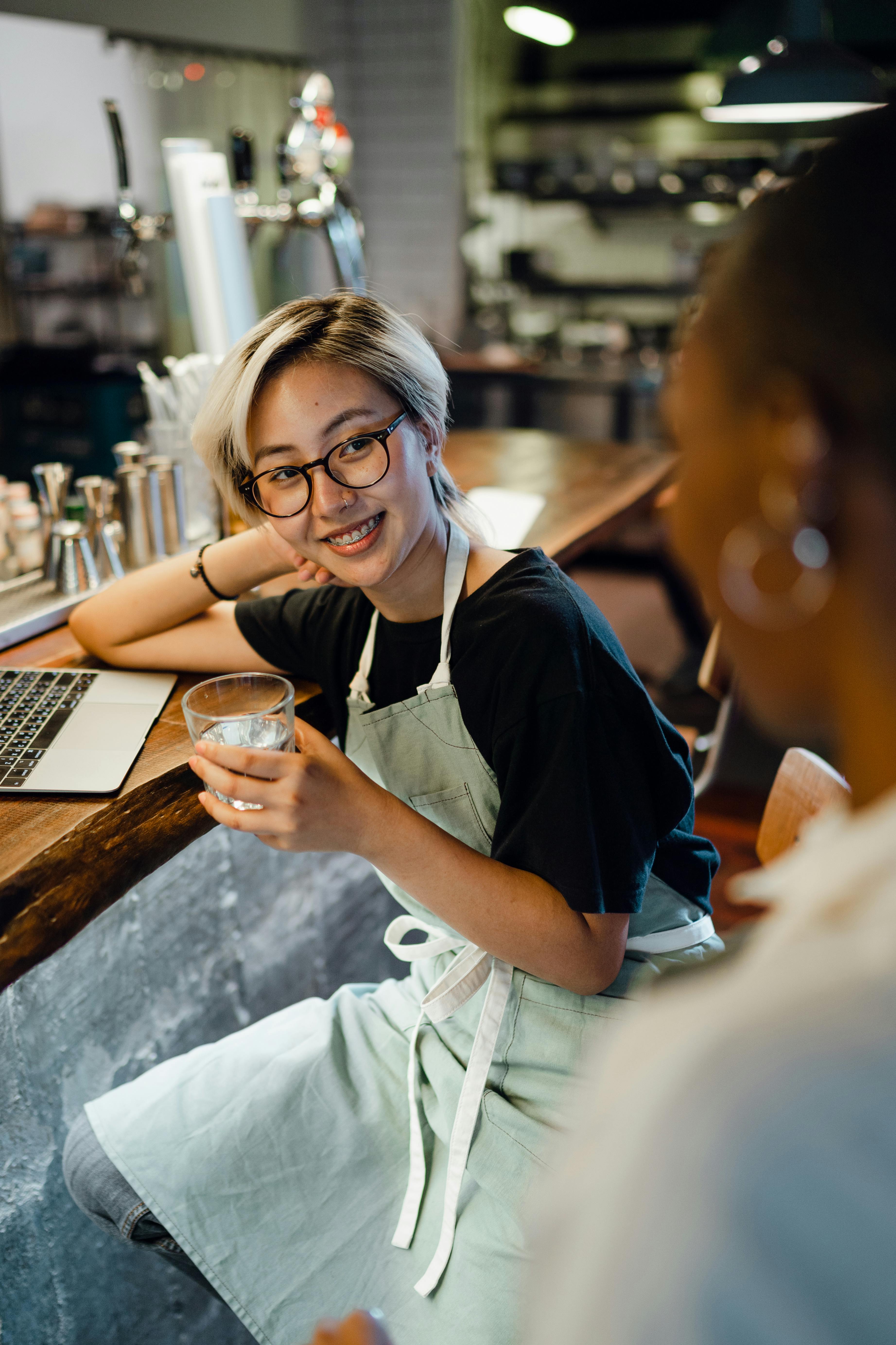 smiling young ethnic woman drinking water and talking to colleague at bar counter
