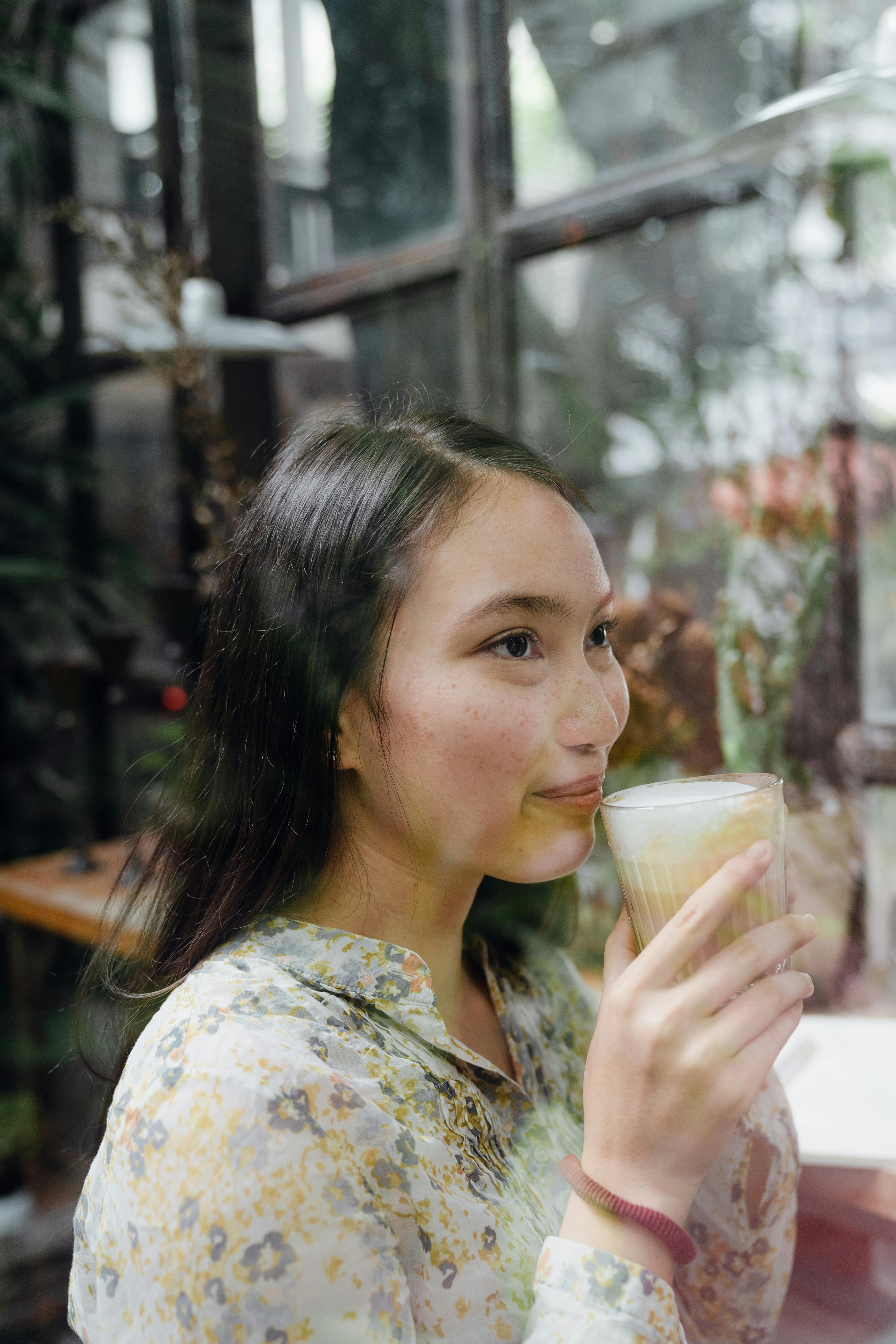 一位穿着花卉衬衫的快乐年轻亚洲女性侧坐在大窗边，手捧一杯芳香的热拿铁，目光梦幻地望向远处