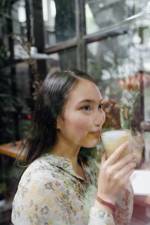 Side view of glad young Asian female in floral blouse sitting with cup of aromatic hot latte against big window and looking away dreamily