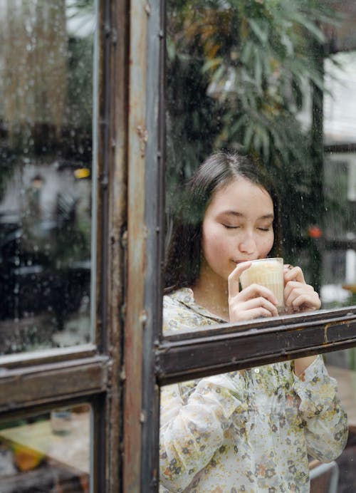 Charming young Asian female in casual clothes standing behind window with eyes closed and enjoying aromatic smell of latte on rainy day