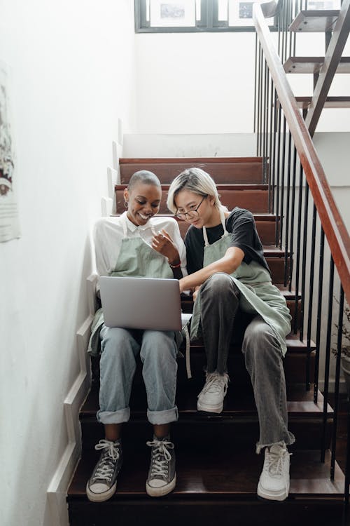 Full body of positive young diverse female friends in trendy clothes and aprons watching funny video on laptop sitting on staircase