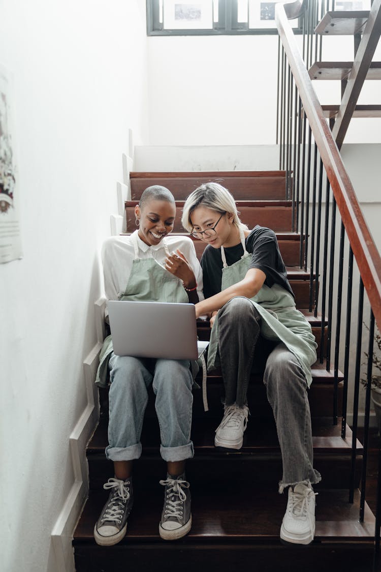 Happy Young Multiethnic Baristas Watching Movie On Laptop Sitting On Stairs During Break