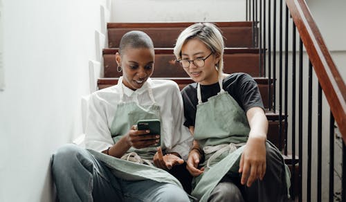 Relaxed young diverse women messaging on smartphone sitting on stairs