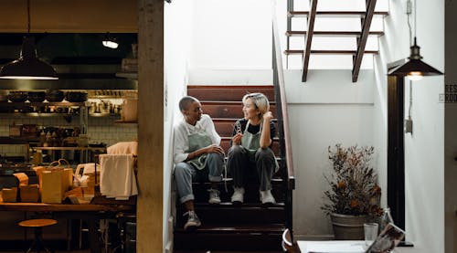 Full length of cheerful young multiracial waitresses in aprons talking to each other while resting on staircase near restaurant kitchen during break