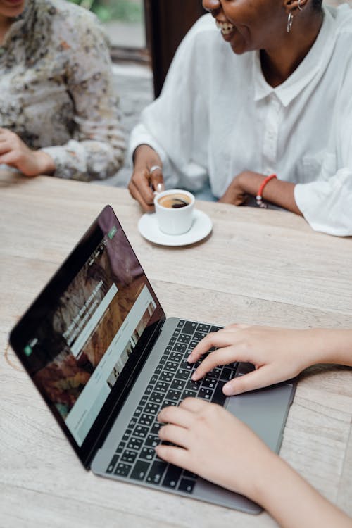 From above of crop anonymous female freelancer typing on laptop keyboard while sitting at table with diverse friends drinking coffee