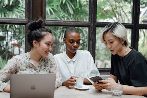 Free Young Asian and African American women checking posts on social media while sitting in cozy cafe with coffee and laptop near large window with exotic green garden outside Stock Photo