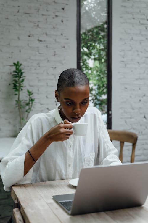 Focused young slim African American business lady with short hair in loose white shirt working on laptop while enjoying small cup of coffee in modern cafe