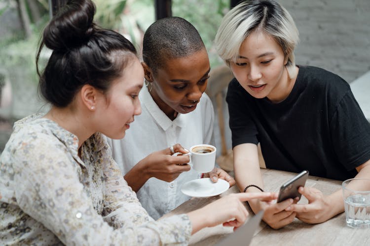 Diverse Female Friends Checking Social Media On Mobile In Cafe