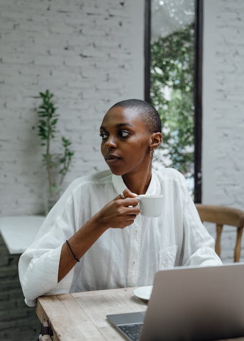 Pensive slender African American female with super short haircut in loose white shirt having small cup of coffee and looking away while working on laptop in modern cafe