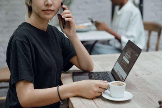 Crop Asian lady in black tee making phone call while sitting at table with coffee and laptop in cafe