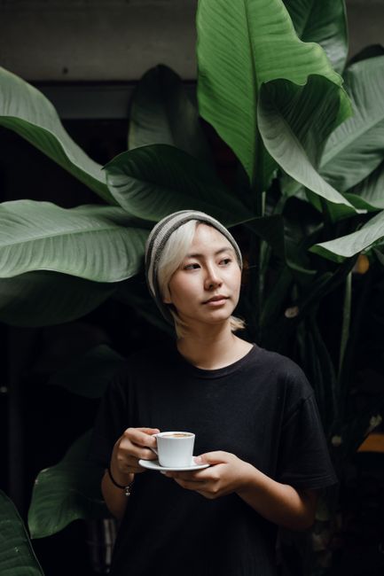 Young slender Asian female in black tee and striped beanie with small cup of coffee in hand standing near plant with large green leaves and looking away in studio