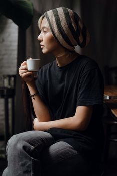 一位穿着牛仔裤、戴着无檐帽的亚洲年轻女性，在黑暗的房间里侧身坐着，一边喝咖啡一边沉思
