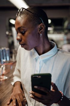 Elegant black woman with super short haircut in white shirt looking at bar counter with mobile in hand