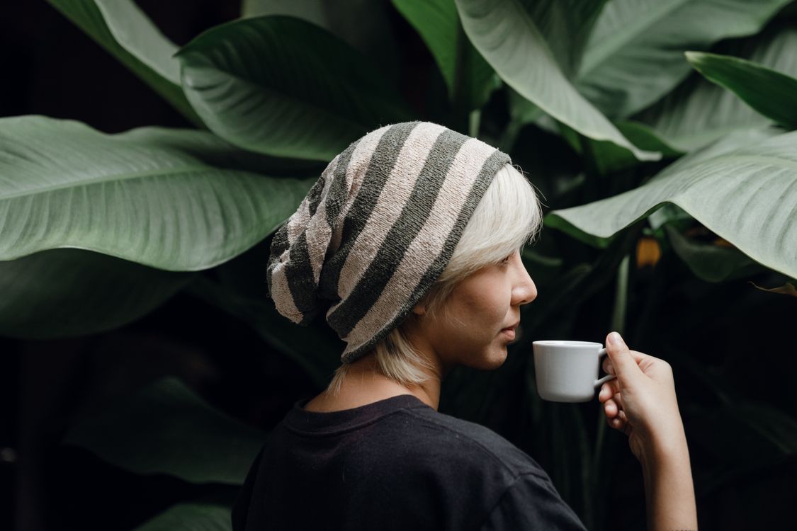 young woman in striped slouchy beanie and black tee with blond fringe drinking cup of aromatic coffee