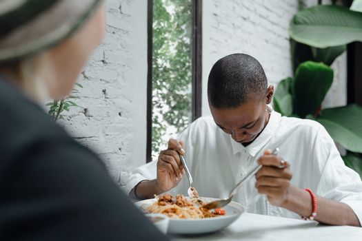 Young black woman looking down at white shirt stained with food while having pasta in cafe together with unrecognizable friend