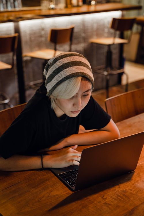 Free From above of young Asian lady with blond bangs in slouchy beanie and black T shirt sitting at table and browsing Internet sites on laptop in modern bar Stock Photo