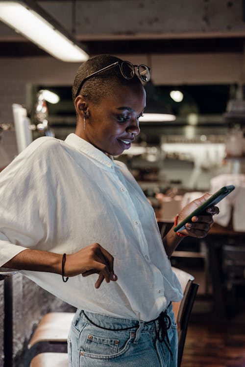 Young confident African American lady in trendy casual outfit with short hairstyle messaging on mobile phone and smiling lightly while leaning on bar counter