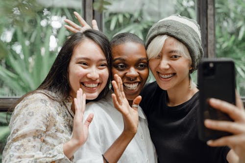 Free Cheerful Asian and African American young ladies waving hands and smiling at phone camera while taking selfie Stock Photo