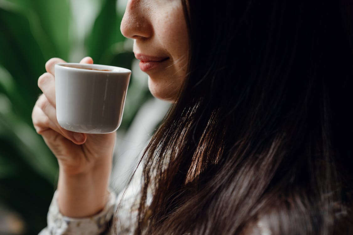 Crop long haired brunette drinking hot aromatic coffee from small ceramic cup in cafe against blurred green pot plants