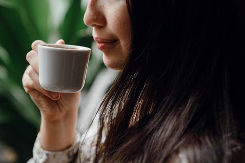 Free Crop long haired brunette drinking hot aromatic coffee from small ceramic cup in cafe against blurred green pot plants Stock Photo
