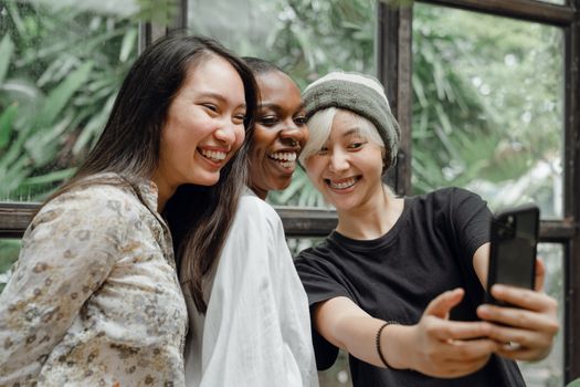 Happy young Asian and African American female friends smiling and taking selfie on smartphone while spending time together in cafe