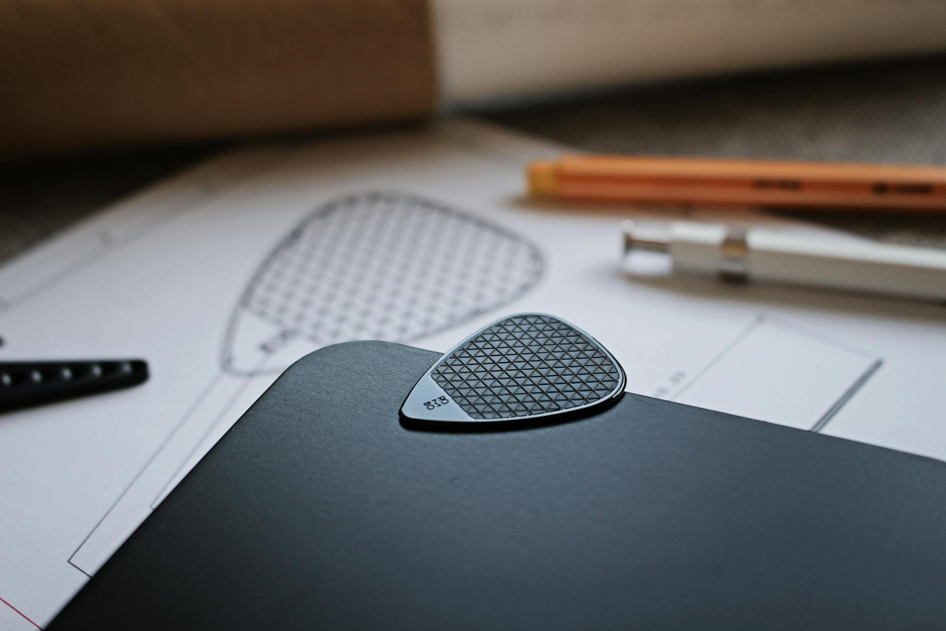 Close-up of a guitar pick on a design blueprint beside drafting tools.