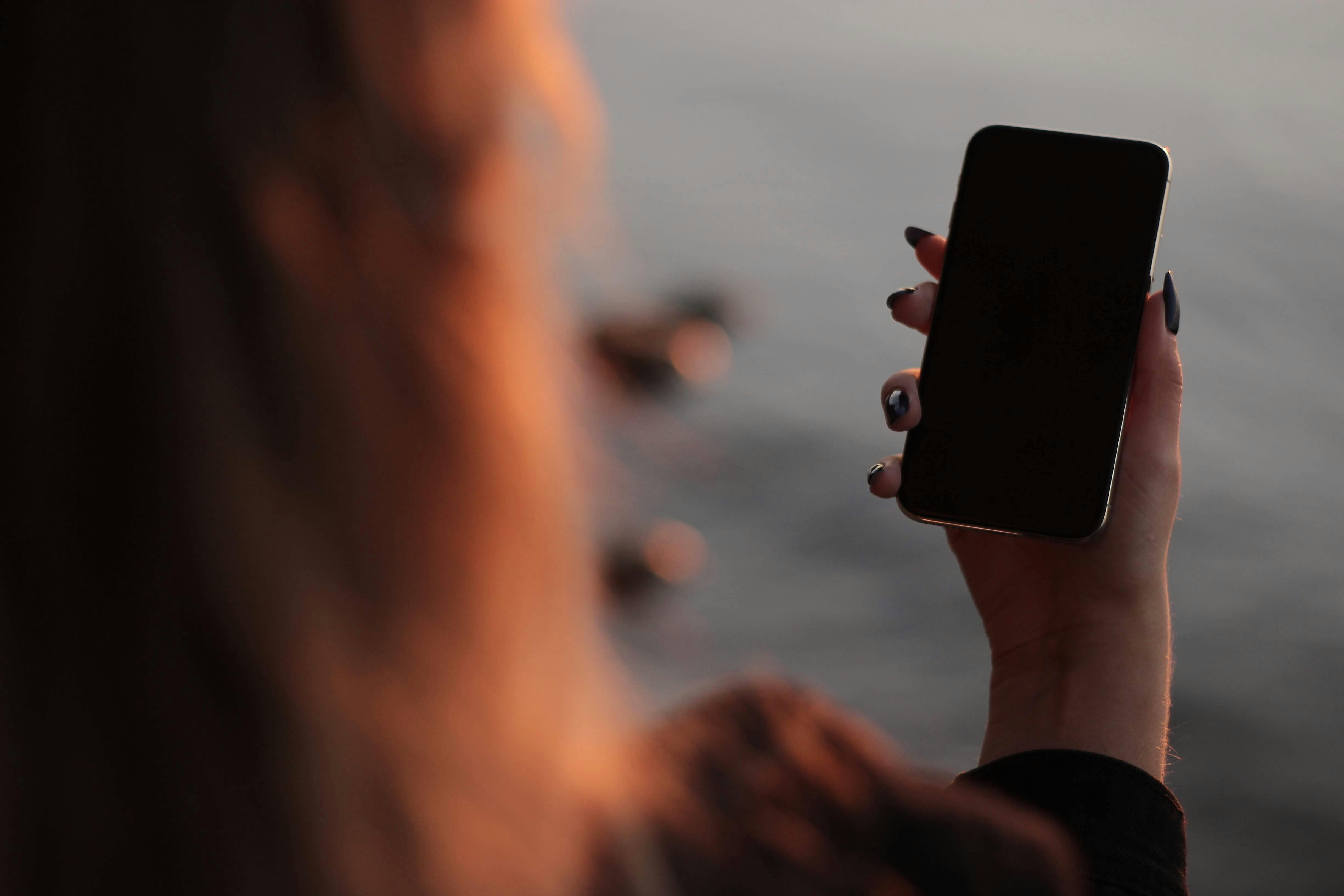 日落时分，一名女子手持智能手机面向水面，营造出宁静而沉思的氛围