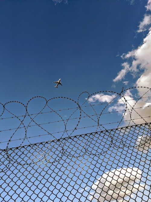 Free Low angle of flying aircraft in bright blue cloudy sky over chain link fence with barb wire Stock Photo