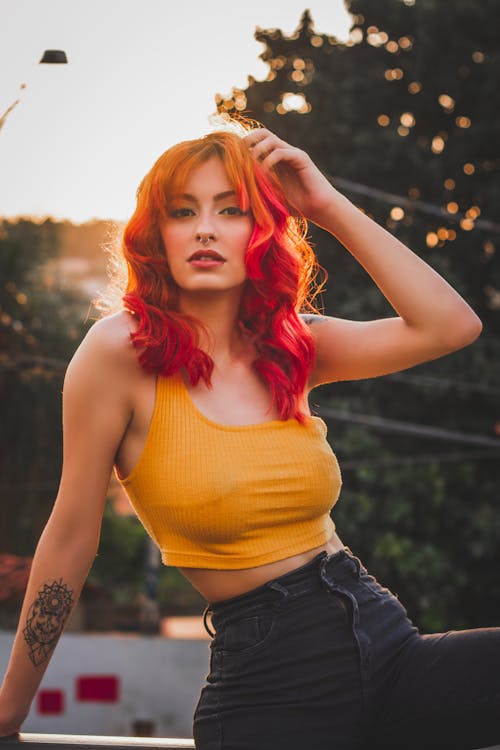 Free Sensual ethnic female with creative tattoo in trendy outfit looking at camera while touching bright red hair in back lit Stock Photo