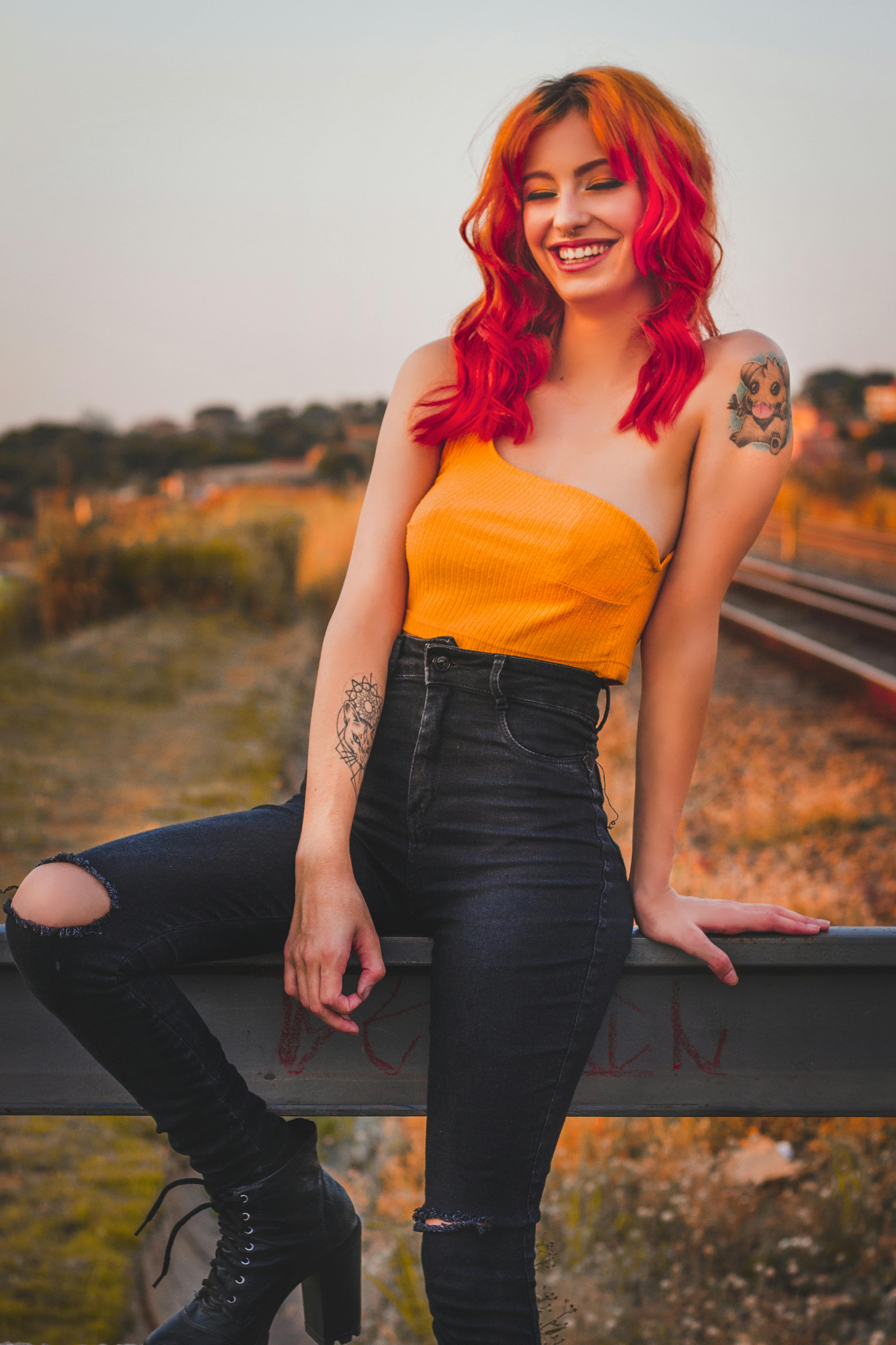 Young slim smiling woman with tattoos and red hair in trendy clothes sitting on concrete fence with closed eyes in countryside