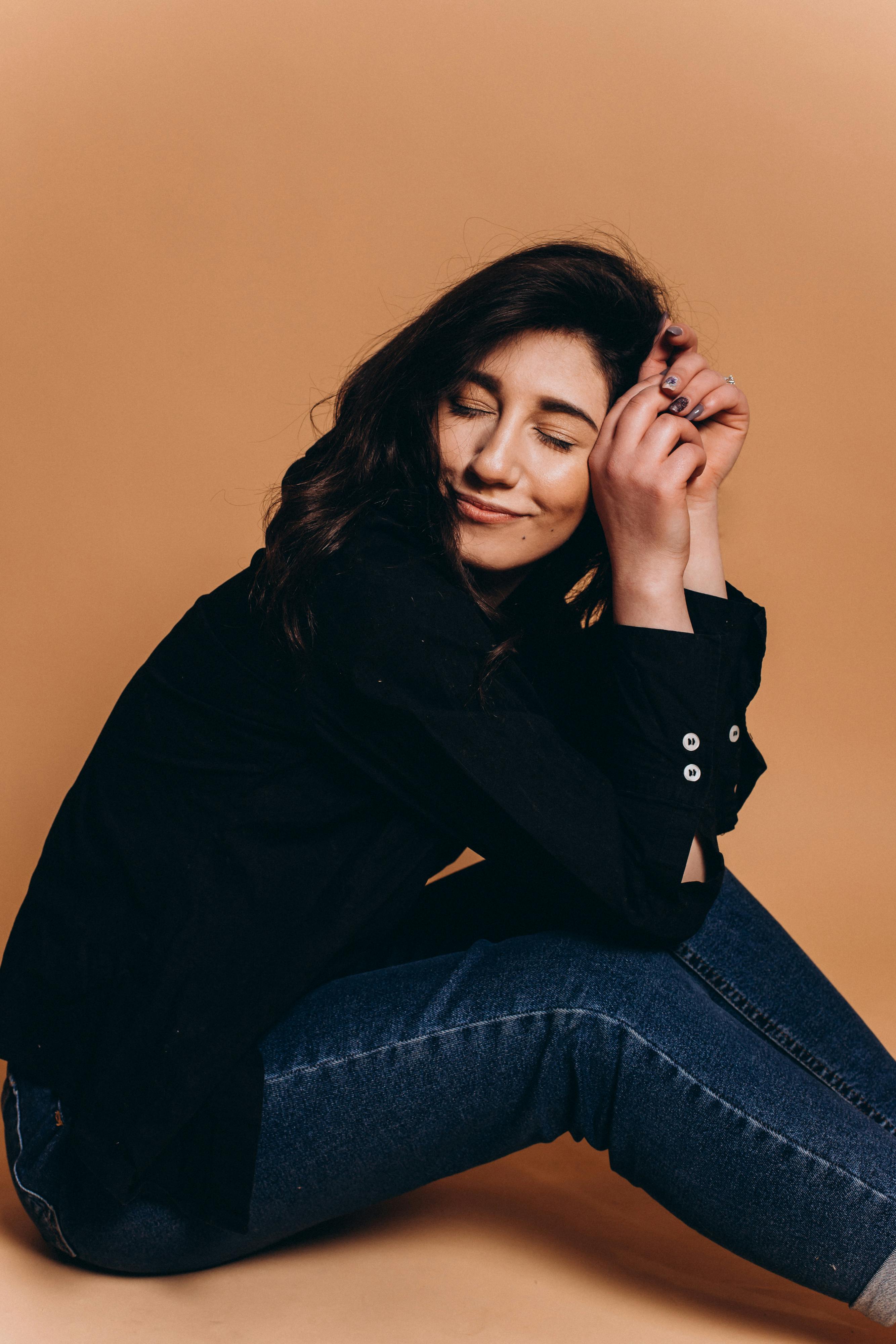 Side view of positive young lady with dark hair relaxing on floor with closed eyes and smiling against beige background