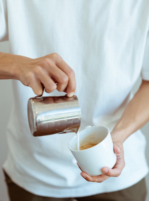 Unrecognizable male barista in white tee pouring fresh whipped milk from metal jug into white ceramic cup while preparing aromatic coffee in modern cafe