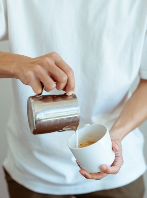 Free Unrecognizable male barista in white tee pouring fresh whipped milk from metal jug into white ceramic cup while preparing aromatic coffee in modern cafe Stock Photo