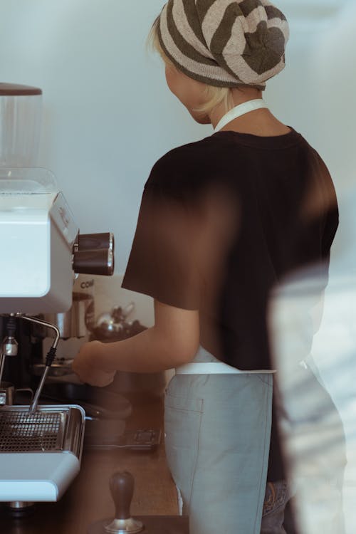 Through glass back view of anonymous female coffee house employee in black T shirt and slouchy knit beanie making fresh coffee using coffee machine