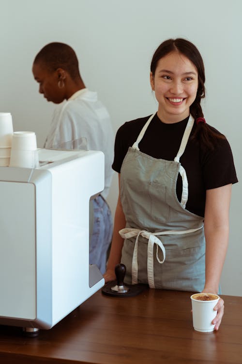 Free Positive Asian female coffee shop worker smiling and serving paper cup of freshly made coffee to customer with African American coworker behind coffeemaker in background Stock Photo