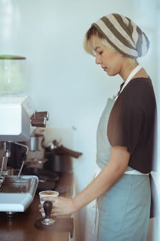Through glass side view of serious young Asian female barista in apron preparing fresh coffee on ordinary workday at cafe