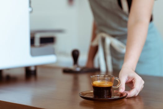 Barista with apron serves a hot espresso in a modern cafe, blurred background.