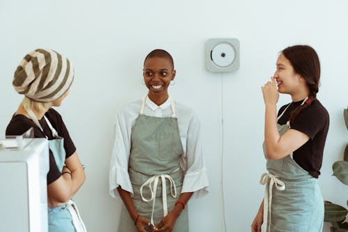 Free Positive young diverse female baristas in apron uniforms communicating with shy newcomer during break at work Stock Photo